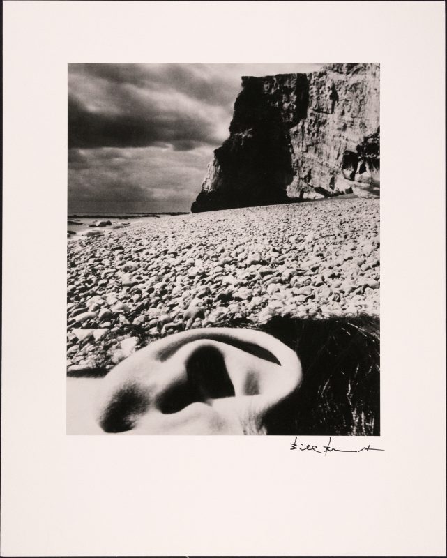 black and white photo of a rocky beach, with a close up of a human ear in front laying down