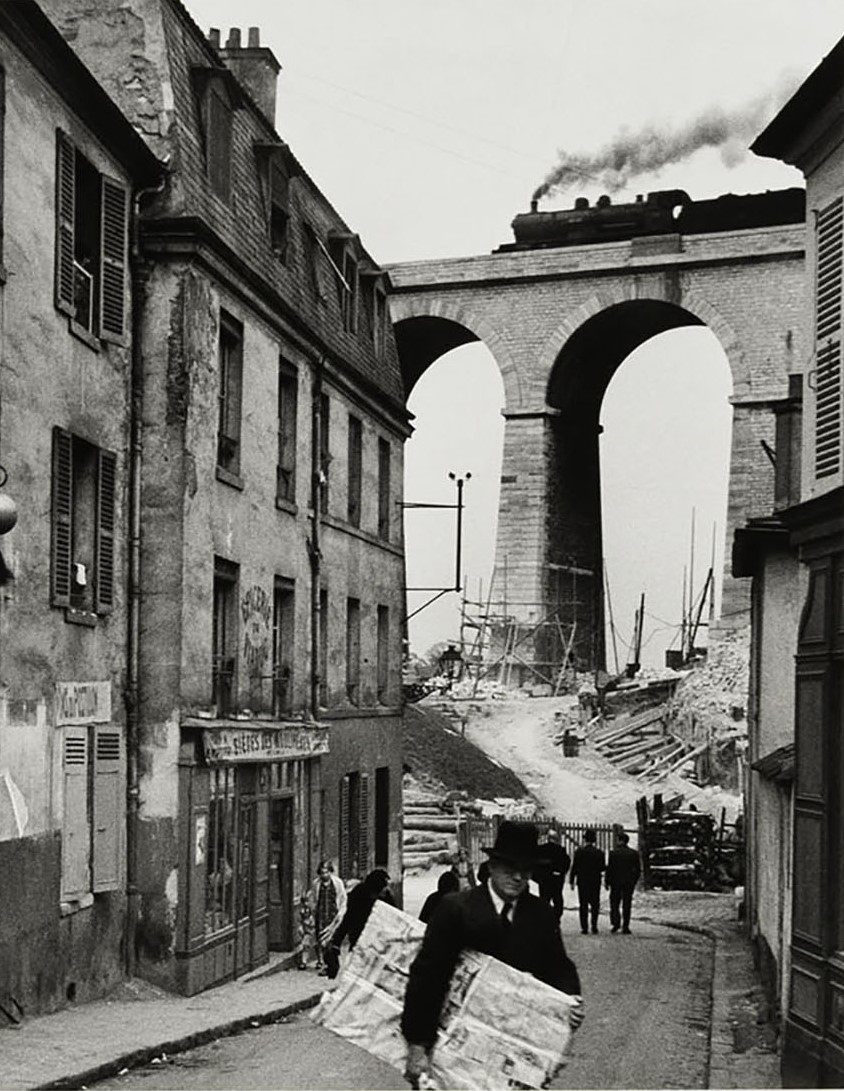 Black and white photo of a street scene with brick buildings on left, a railroad bridge behind, and a person walking across the street carrying a parcel