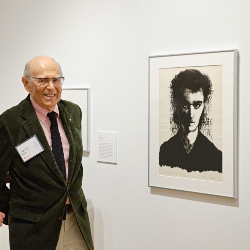 Interior view of a gallery with a man standing in a dark suit jacket, tie, and pink dress shirt next to a black and white portrait framed on the wall