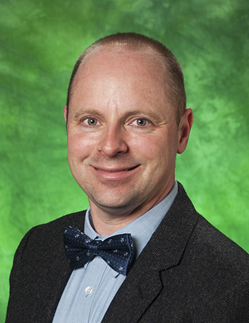 Color photograph of Clark A. Pomerleau, a clean-shaven white man in his forties with short-cropped brown hair and smiling hazel eyes. He is dressed in a light blue button-down shirt, navy bowtie, and tweedy grey suit jacket. The photo's background is green.