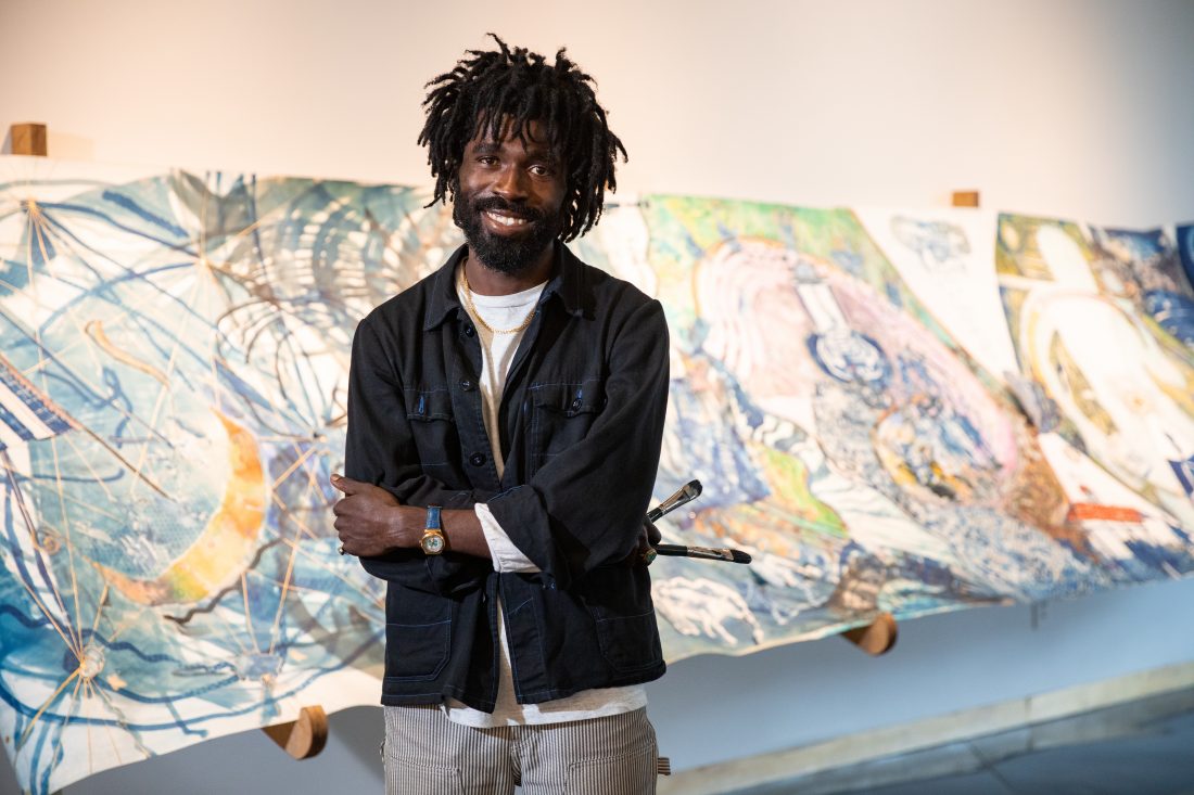 Black male with a blazer on, crossed arms, standing in front of a white wall with a handprinted scroll behind