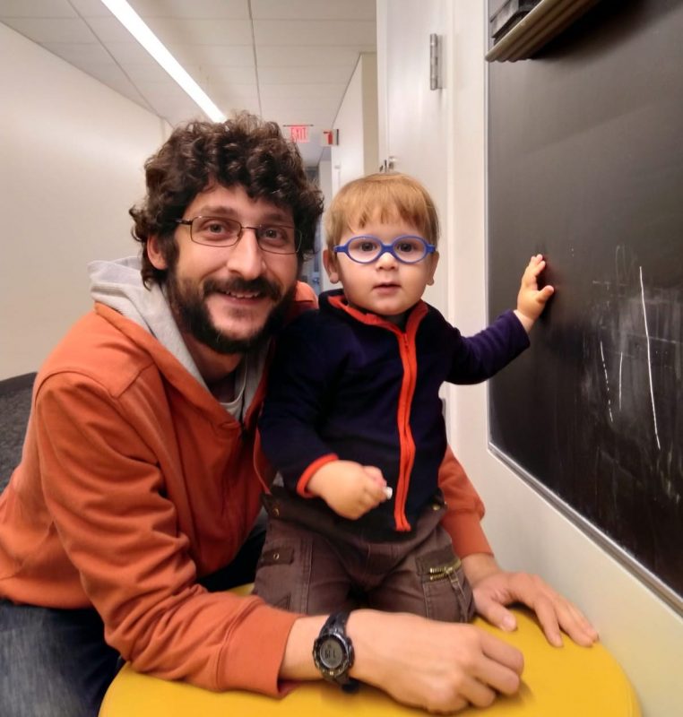 A man in an orange hoodie poses with a toddler in front of a black board