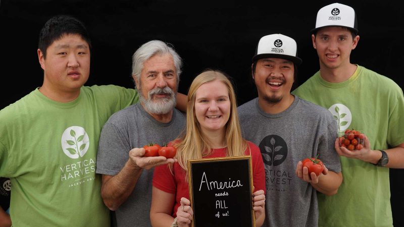 five people holding tomatoes looking into camera. the center person is holding a sign that says america needs all of us