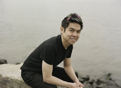Duy Đoàn smiles while sitting on a rock near a body of water.