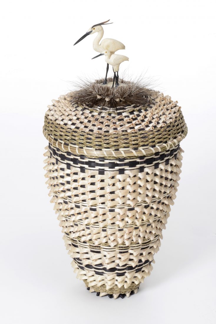 Tall woven basket in light tan and brown colors, top of lid has feather like weavings and a carved bird (egret) made of stone
