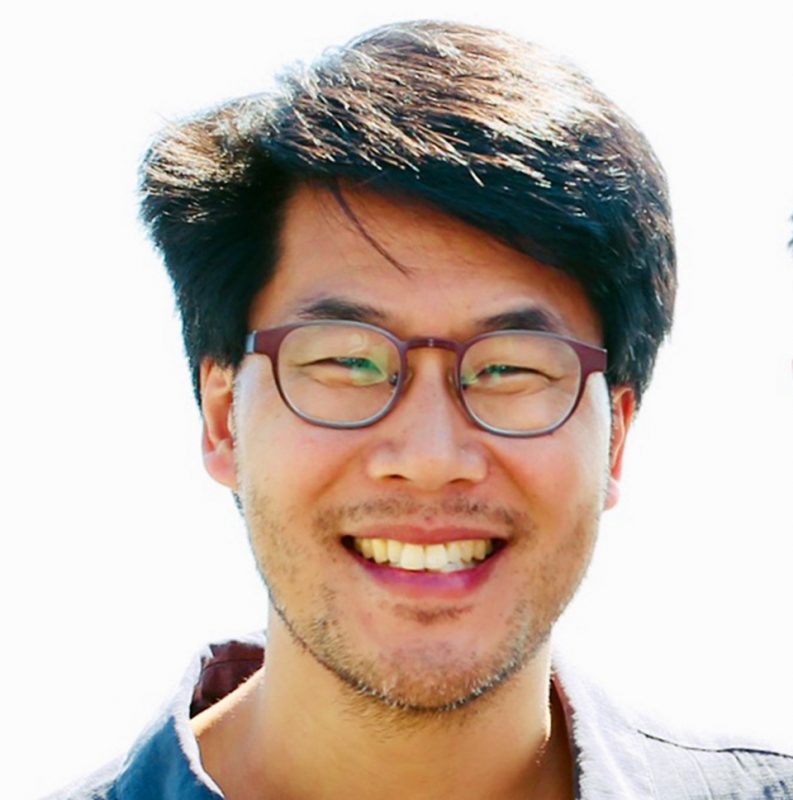 A close-up color photo of Dr. Eric Lai smiling for the camera against a bright white background. Dr. Lai has dark hair and wears glasses and a blue T-shirt. 