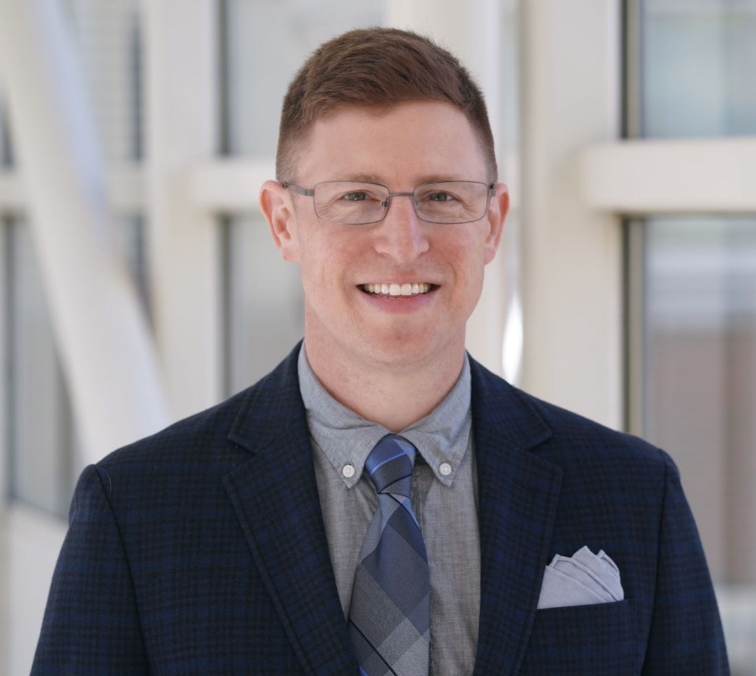 A color photo of Dr. Michael Feigin smiling toward the camera. He has short hair, wears glasses and a dark blue suit with a lighter blue shirt, tie, and pocket square. He stands in front of large, white-framed windows of a building. 