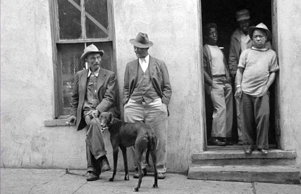 black and white photo of Black gentlemen and a dog assembled outside of a concrete building and in an open doorway