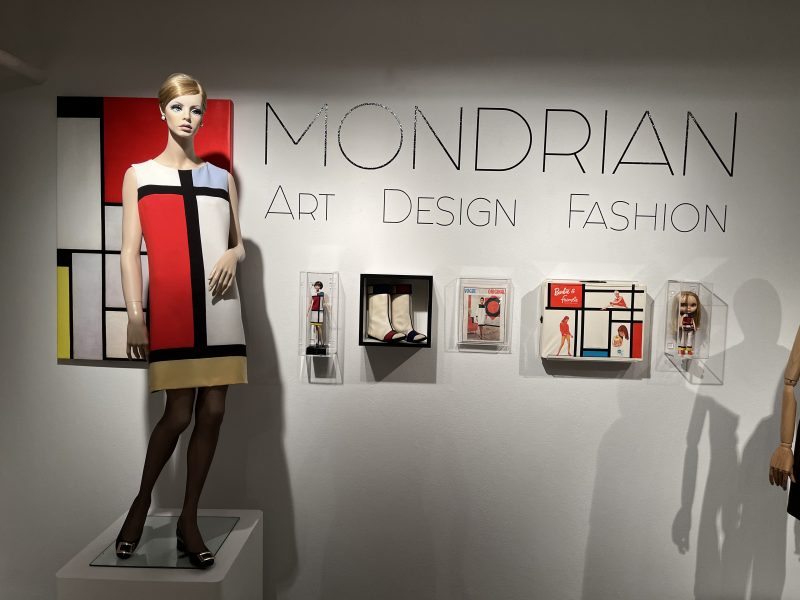 A gallery with a model wearing a dress and objects displayed on the wall. The wall has text that says Mondrian Art Design Fashion