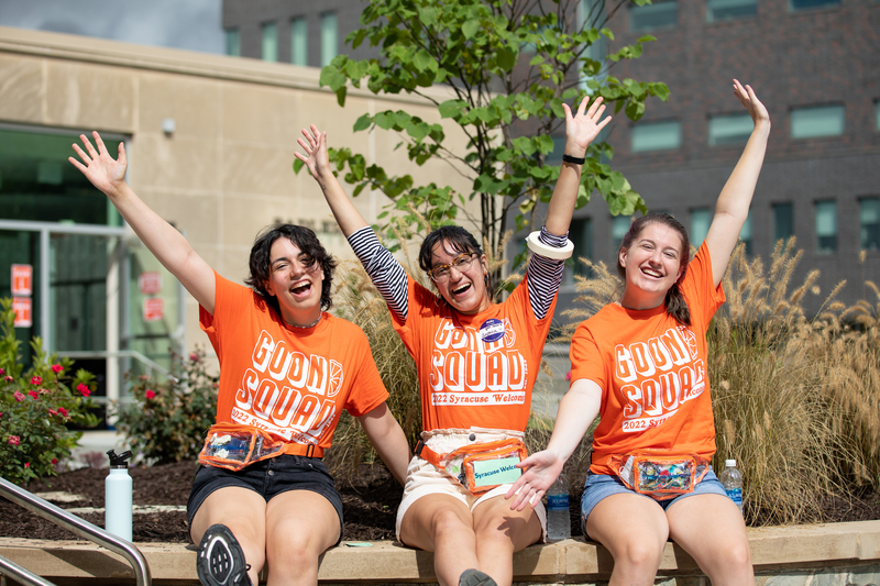 Three students volunteering for Goon Squad sit on a wall outside Sadler Hall and smile for a photo