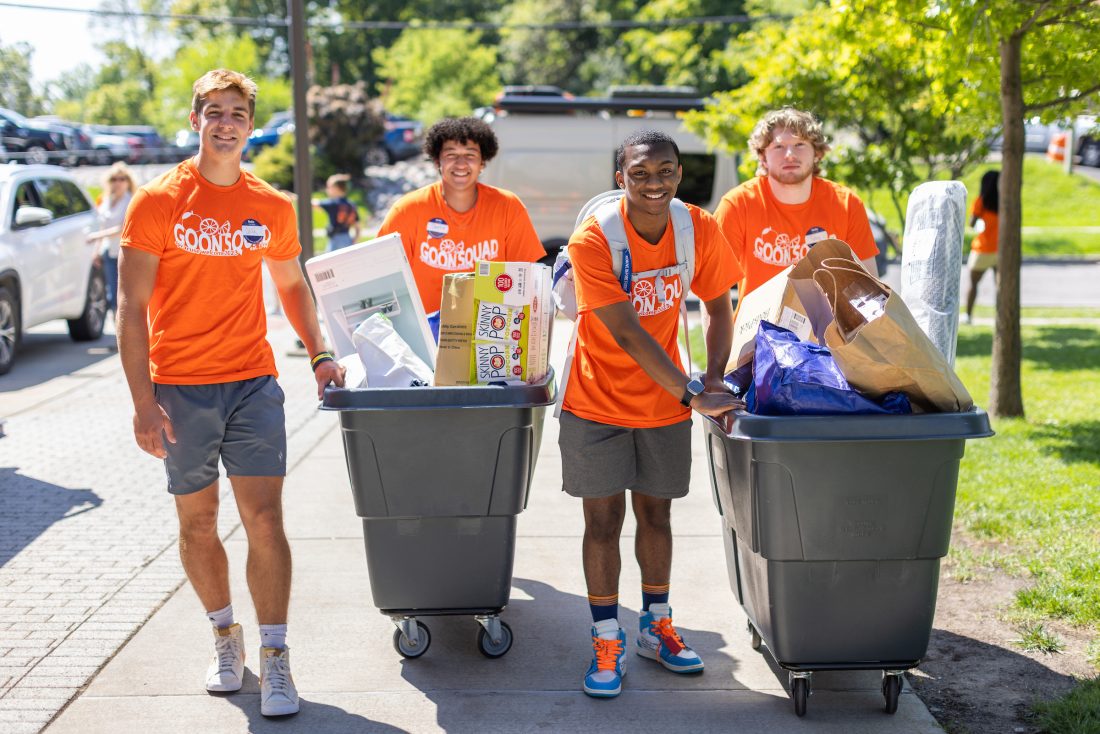Four students moving bins with items on a sidewalk.