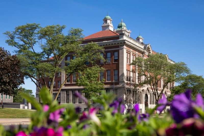 Lyman Hall at Syracuse University on a sunny day. The historic brick building features tall windows, ornate architectural details, and green domed towers on the roof. Trees partially frame the building, with vibrant purple and pink flowers in the foreground under a clear blue sky.