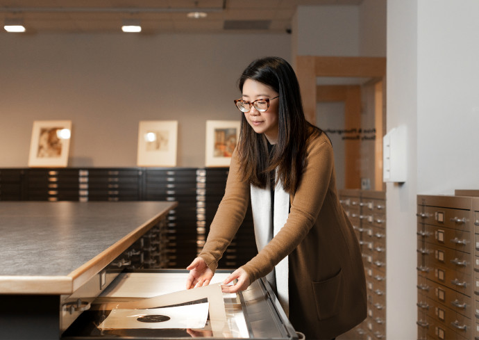 Woman wearing a brown sweater with long dark hair and glasses, placing artowrk in a large metal file cabinet