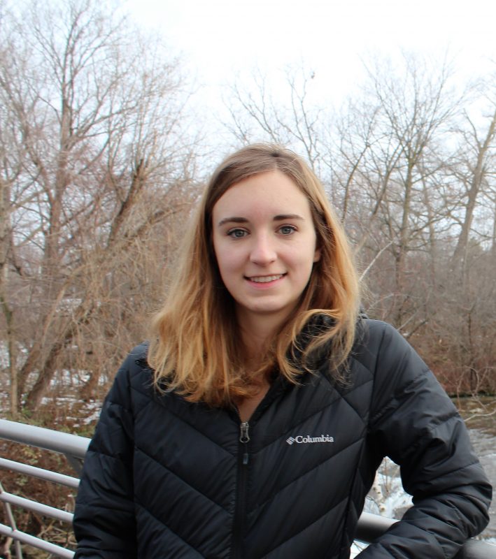 A color photo of Mariah Maxwell smiling toward the camera. She wears a black winter coat and stands outside in front of a creek or river. There are leaf-bare trees in the background. 