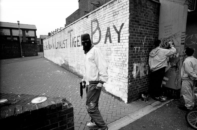 Belfast, Northern Ireland, 1990 A hooded paramilitary gunman stands in front of a painted wall that reads “You Are Now In Loyalist Tiger Bay.”