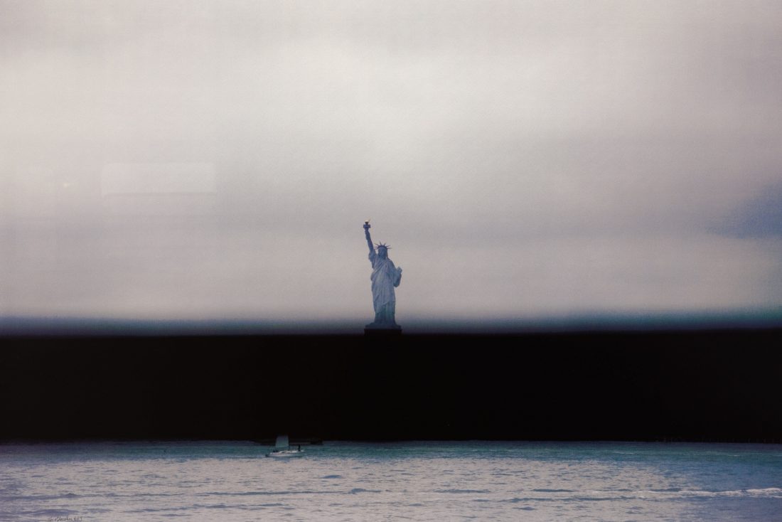Hazy fine art photo of the statue of liberty from a distance, in the middle of the photo with a body of water in front, and a dark line in the middle.