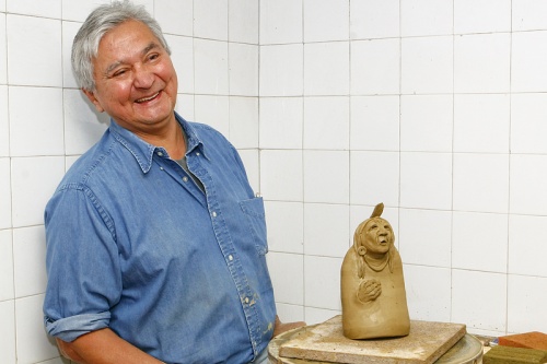 Interior image of a male figure, at the left, leaning against a wall, wearing a button up blue denim shirt, with a clay figure carving on a table to his right.
