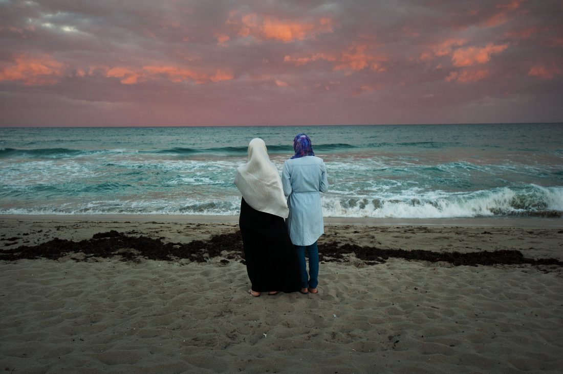 two people stand on a beach with their backs turned to the camera.