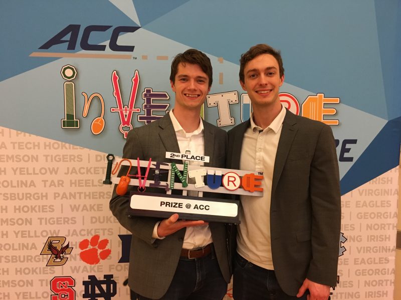 two young men holding InVenture prize with ACC InVenture banner in background