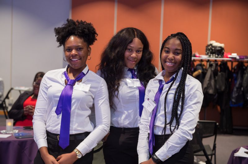 A group of smiling Dimensions mentors and mentees gather on campus in white button-up shirts with purple ties.