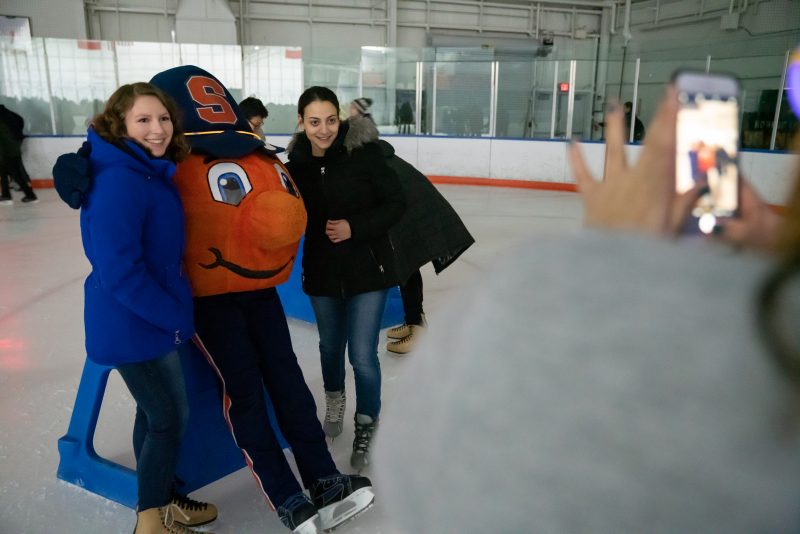 A student takes a photo of their friends on the ice rink posing with Otto the Orange during Late Night at the Rink.