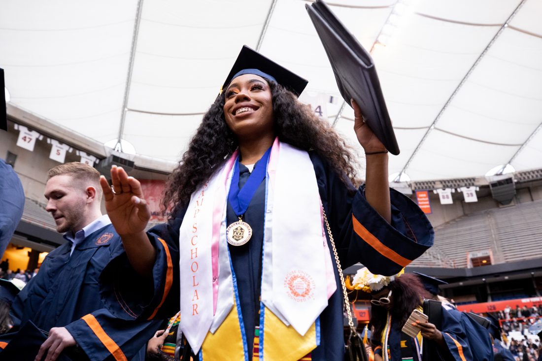 Wearing a cap, gown and received honors, a Syracuse University student stands and smiles at graduation in the JMA Wireless Dome while holding a padfolio.