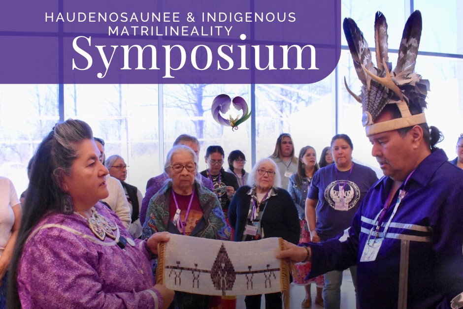 woman holding wampum belt on left, man wearing feather headdress on right, people in background with symposium title at top