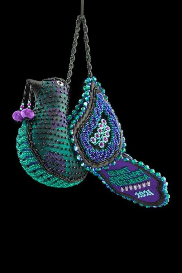 Woven and beaded bird with dark teal and purple coloring , a beaded cherry handing from his mouth, and sparkling beads across its surface
