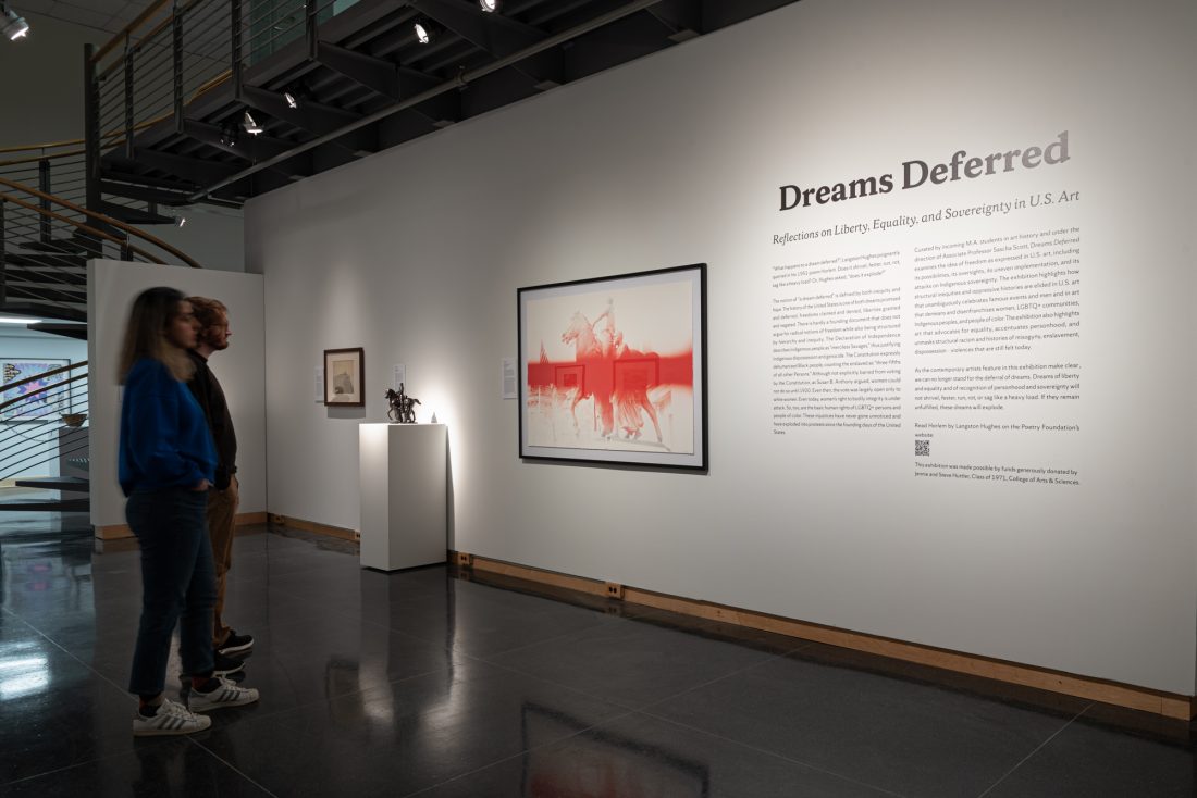 Interior of a museum with artwork installed on wall and two figured standing on left