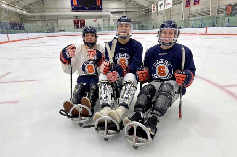 Three members of the Syracuse University Men's Club Hockey sit on sled hockey sleds and hold sled hockey gear on the Tennity Pavilion ice.