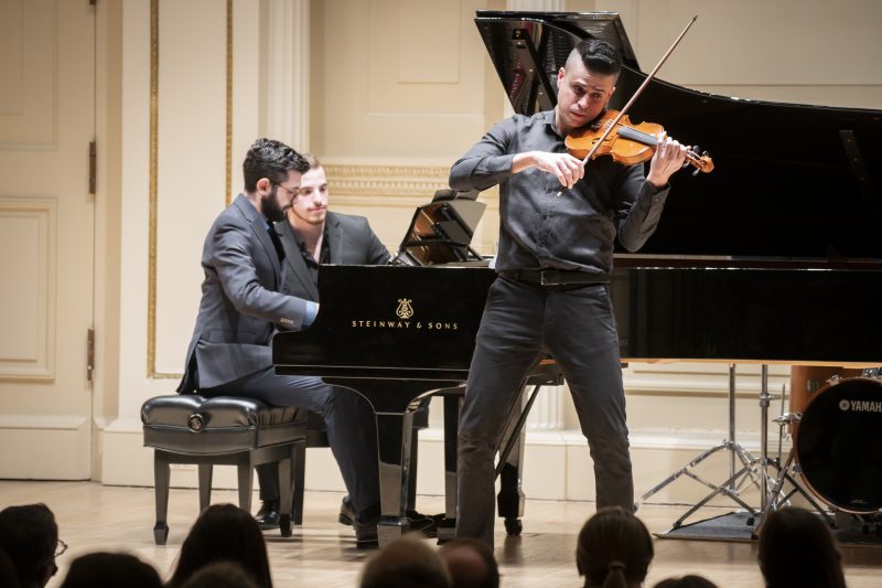 A violinist plays on a stage with a pianist playing piano behind him