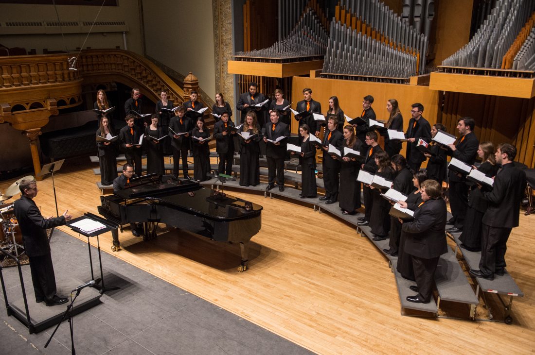 A group of singers stand on risers on a stage with a piano and conductor