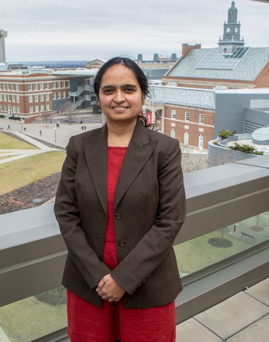 Dr. Shailaja Paik, a South Asian woman wearing a dark brown blazer with a red undershirt. She is standing in front of several buildings. She is smiling and her arms are crossed over in front of her. 