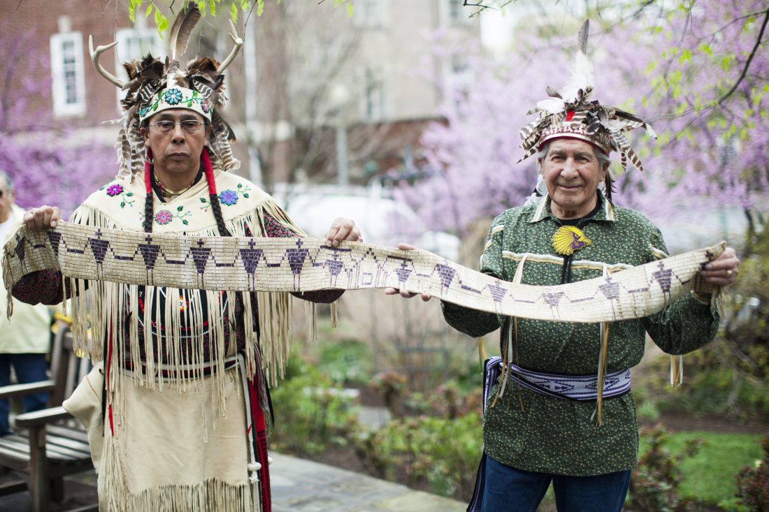 Two indigenous males standing in traditional clothing holding a long wampum beaded work in their hands