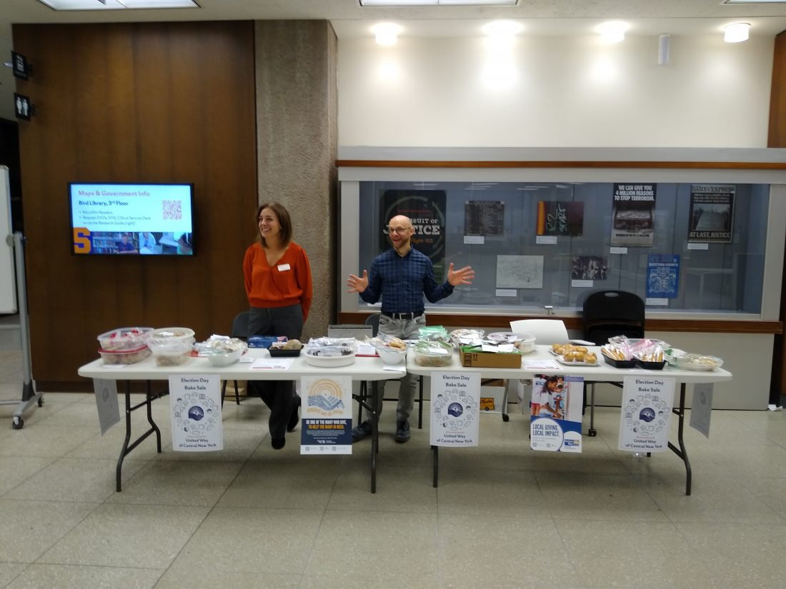 2 people standing behind table of baked goods