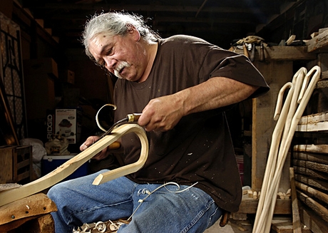 Man with gray hair pulled back, wearing blue jean pants, a black long sleeved shirt. Sitting on a chair, carving a rounded wooden stick