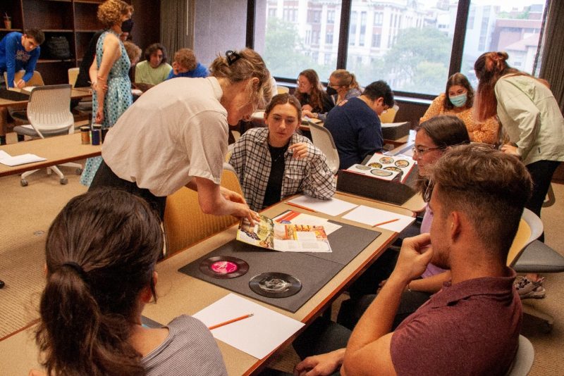 person standing with book and pointing at page while students sitting in chairs hve records in front of them