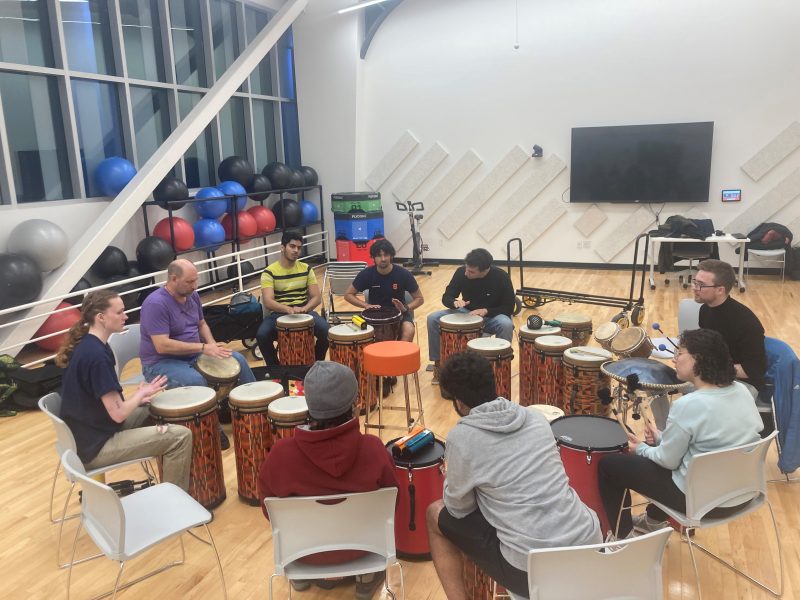 People Drumming with hand drums and percussion instruments in a seated position within a fitness room in the Barnes Center