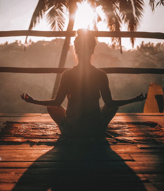 A picture of a woman meditating on a wooden porch facing a tropical sunrise.
