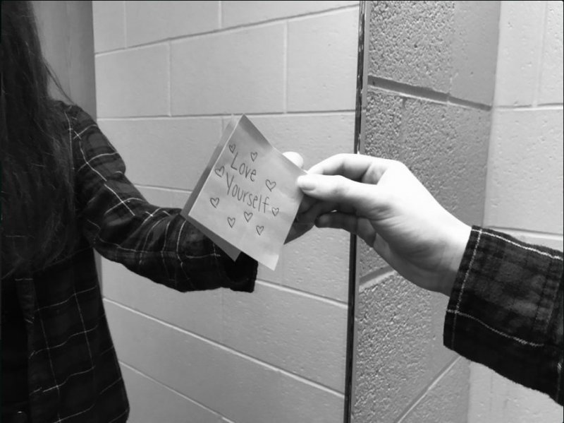 A person (faceless) standing in front of a mirror touching a sticky note displayed on the mirror that says 