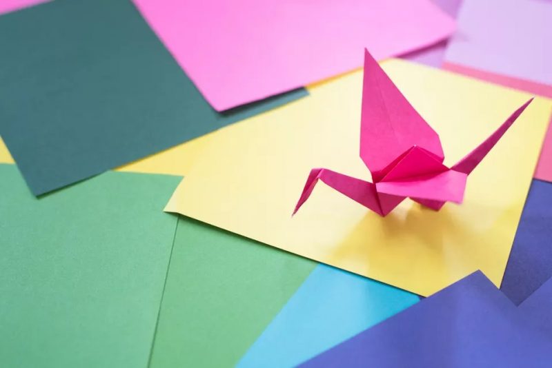 Numerous sheets of paper of various colors including blue, yellow, greens and pink covering a table surface with a pink origami crane on top of a yellow sheet