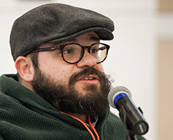 Color photograph of Prof. Joseph A. Stramando, a cis-gender, white man with dwarfism. He is wearing a green sweater, round framed eyeglasses, a dark grey cap and a neon orange lanyard around his neck. He has a thick beard of dark brown hair and is speaking into a microphone while gesturing with his right hand, in which he is holding a clicker for a slide presentation.