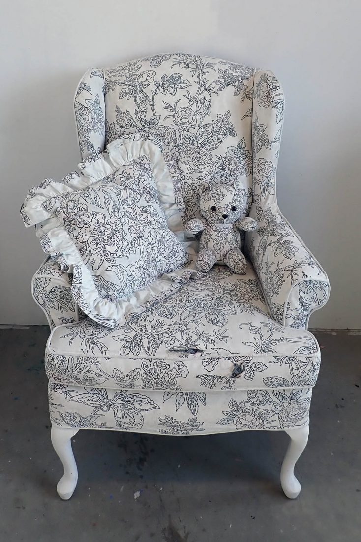 Armchair with a white background with hand drawn black designs. A decorative pillow and a small teddy bear in the same design is perched on the chair 